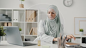 Portrait of muslim woman coming to office sanitizing hands with gel then working at desk