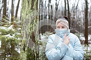 Portrait of mysterious middle-aged woman covering face with blue warm puffy jacket, standing in forest in snowy winter.