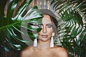 Portrait of naked brunette model girl with amazing blue eyes and stylish earrings, looks through the big green leaves
