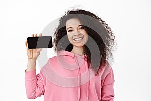 Portrait of natural smiling girl showing horizontal smartphone screen, online shopping app or video game on moible phone