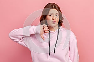 Portrait of naughty dissatisfied brunette teen in hoodie frowning angrily and showing thumbs down gesture, expressing disapproval