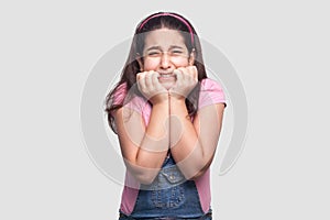 Portrait of nervous sad alonel brunette young girl in pink t-shirt and blue denim overalls standing with worry face and bitting