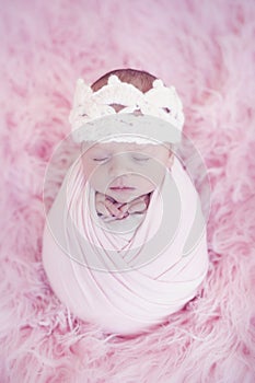 Portrait of newborn baby girl wrapped in pink wrap with knitted woolen crown headband. Infant on furry fluffy background