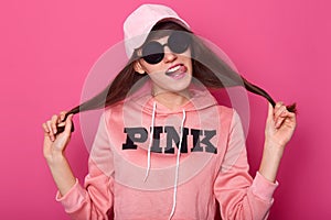 Portrait of nice cute attractive girl pulling her hair asides, wears shortened hoodie, cap and stylish black sunglasses, shows her