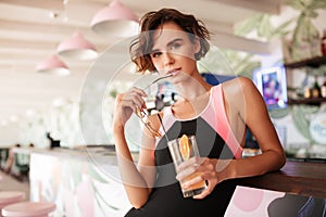 Portrait of nice girl in trendy swimsuit leaning on beach bar counter with sunglasses and cocktail in hands. Young