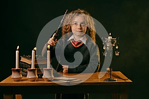 Little witch dressed in dark clothes sitting at the table against a black smoky background. There are magic wand, books