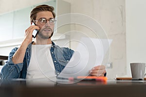 Smiling man, he is sitting at a table, talking on the phone with his laptop, notebook and glasses in front of him