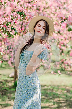 Portrait of nice stylish young girl in straw hat in front of blooming pink apple garden