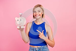 Portrait of nice young girl direct finger money bank pig wear blue top isolated on pink color background