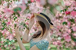 Portrait of nice young girl holding straw hat, in front of blooming pink apple garden, profile view