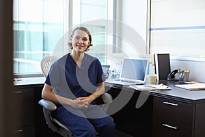 Portrait Of Nurse Wearing Scrubs Sitting At Desk In Office