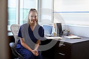 Portrait Of Nurse Wearing Scrubs Sitting At Desk In Office