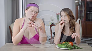 Portrait of obese Caucasian woman eating sweet delicious chocolate as slim arrogant lady chewing green salad leaves