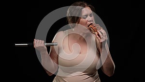 Portrait of obese Caucasian woman enjoying taste of unhealthy sweet bun lifting dumbbell at black background. Overweight