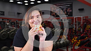 Portrait of obese woman in gym eating hamburger smiling and looking at camera. Food temptation. Overweight concept