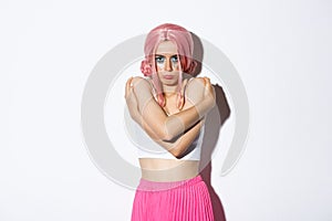 Portrait of offended cute girl hugging herself and sulking, feeling insulted or sad about something, wearing pink wig