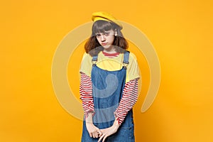 Portrait of offended disconcerted girl teenager in french beret, denim sundress standing isolated on yellow wall