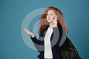 Portrait og beautiful teen redhead girl with wavy hair and freckles talking on phone with cynical face, arguing with her