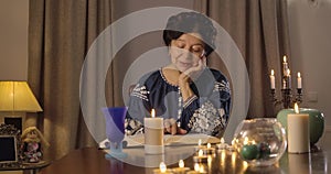 Portrait of old Caucasian brunette woman sitting at the table with lighting candles and reading. Clairvoyant predicting