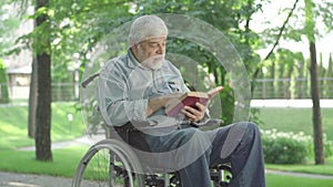 Portrait of old Caucasian man in wheelchair reading book out loud in sunny summer park. Senior disabled retiree turning