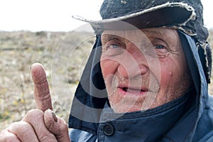 Portrait of an old man in a messy clothes and hat who raised his index finger, I have an idea
