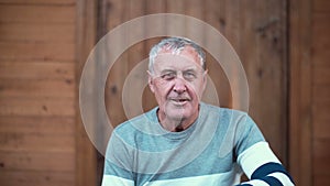 Portrait of old man sitting on the porch of the house. Pensioner looking to the camera, smiling and smoking. 4K