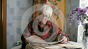 Portrait of an old woman in glasses reading a newspaper while sitting at a table at home