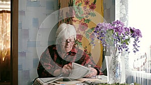 Portrait of an old woman in glasses reading a newspaper while sitting at a table at home