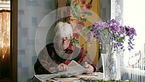 Portrait of an old woman in glasses reading a newspaper while sitting at a table at home