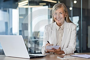 Portrait of an older gray-haired happy business woman working in the office. He sits with a laptop in a suit, holds a