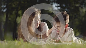 Portrait of older sister spending time with younger brother reading book or magazine outdoors. The boy and girl lying on