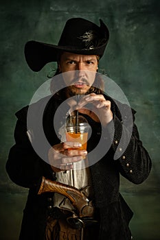 Portrait of one brutal man, medeival pirate with pistol drinking orange juice isolated over dark background.