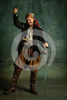 Portrait of one brutal man, medeival pirate raising bottle of rum isolated over dark background.