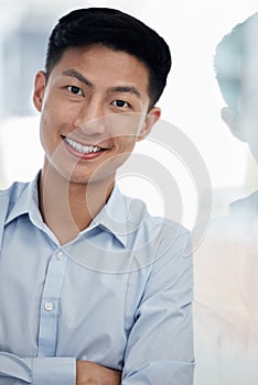 Portrait of one confident young asian business man standing with arms crossed against a glass wall in an office. Happy