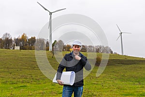 Portrait of one male engineer holding a plan at wind turbine at autumn cloudy sky green meadow