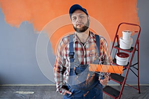 Portrait of one male house painter worker in blue overalls and cap with painting roller before orange wall.
