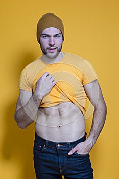 Portrait of One Positive Caucasian Handsome Man In Yellow Tshirt Posing in Warm Hat Over Yellow Background While Pulling Shirt Off