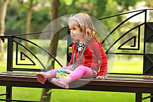 Portrait of a one year old cute little curly girl wearing red cardigan and a headband with a bowl sitting on a bench in the park