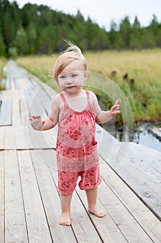 Portrait of a one-year-old girl standing on a wooden footbridge on a pier by the river