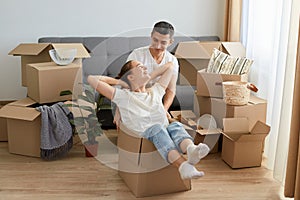 Portrait of optimistic couple having fun with cardboard boxes in new flat at moving day, wife and husband rejoicing to relocate