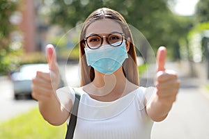Portrait of optimistic girl wearing protective mask showing thumbs up in city street