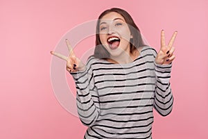 Portrait of optimistic happy woman in striped sweatshirt showing double victory gesture, peace hand sign