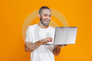 Portrait of optimistic man 30s in white t-shirt holding silver laptop, isolated over yellow background