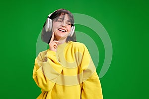 Portrait of overjoyed, young woman listening music in headphones and smiling against vibrant green studio background.