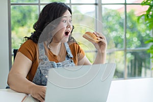 Portrait of overweight asian woman sitting and holding a hamburger open her mouth and hesitate of eating while using a laptop
