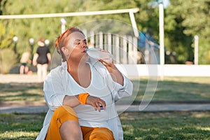 Portrait of an overweight woman sits on the grass in a Park and drinks water from a bottle. Thirst and rest after a walk