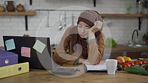 Portrait of overwhelmed young slim beautiful woman in hijab falling asleep sitting in kitchen. Tired exhausted Middle