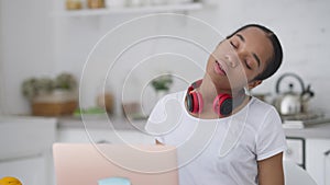 Portrait of overworked young slim woman stretching spasm neck sitting at kitchen table in home office. Beautiful African