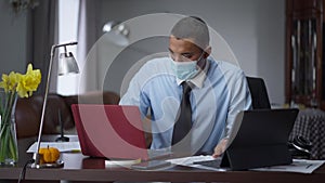 Portrait of overworking African American man in Covid face mask typing on laptop and tablet keyboards, holding head in