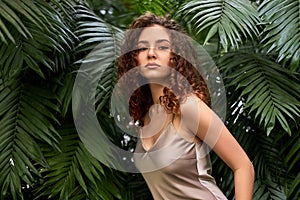 portrait of pacified curly woman posing against backdrop of tropical plant